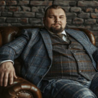 A full-body photo of an overweight man with a short beard, wearing a blue and brown plaid three-piece suit sitting in a leather chair against a dark brick wall. He is looking at the camera with a confident smile on his face. Studio lighting, photorealistic, shot in the style of canon eos r5. --ar 3:2 --style raw Job ID: 067ac235-cce0-435e-821a-8d11a1dee649